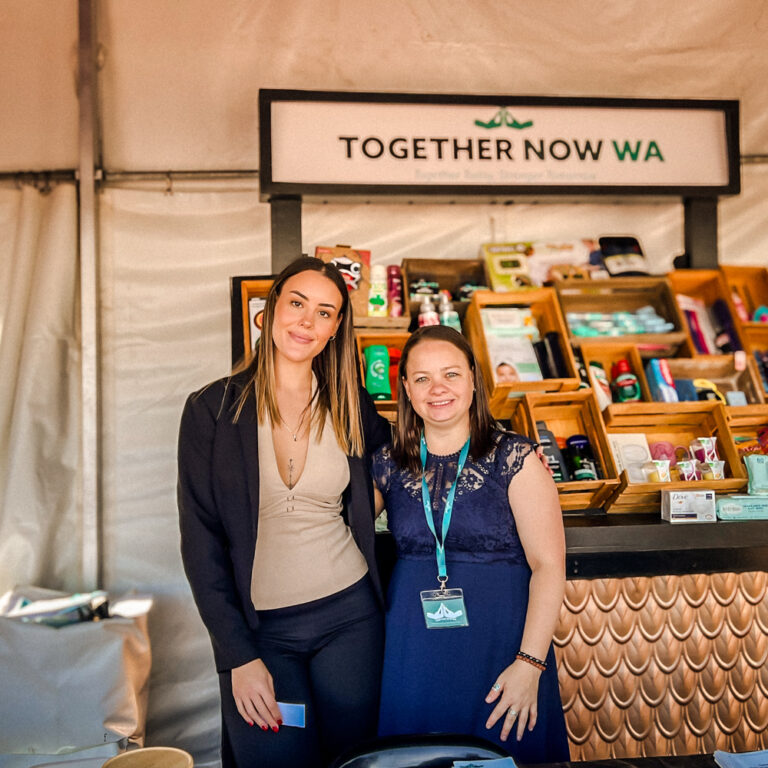 Two women smiling in front of the Together Now WA stall at a community event. Behind them is a display of personal care items and resources in wooden compartments under a branded sign that reads "TOGETHER NOW WA."