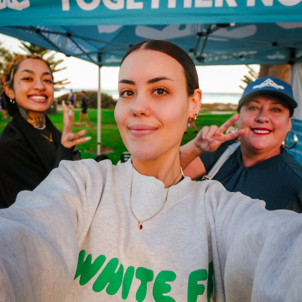 Three women smile for a selfie in front of a blue Together Now WA marquee at an outdoor event. The woman in front wears a light grey sweatshirt with green lettering, while the two behind her flash peace signs and grin warmly. A grassy park and ocean can be seen in the background.