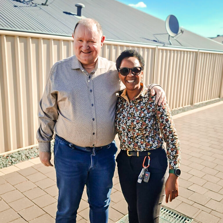 Two people smiling with arms around each other outside a home. One wears a patterned shirt and jeans, the other wears a floral button-up with sunglasses and a keychain on her belt. They stand on a paved pathway with a fence and satellite dish in the background.