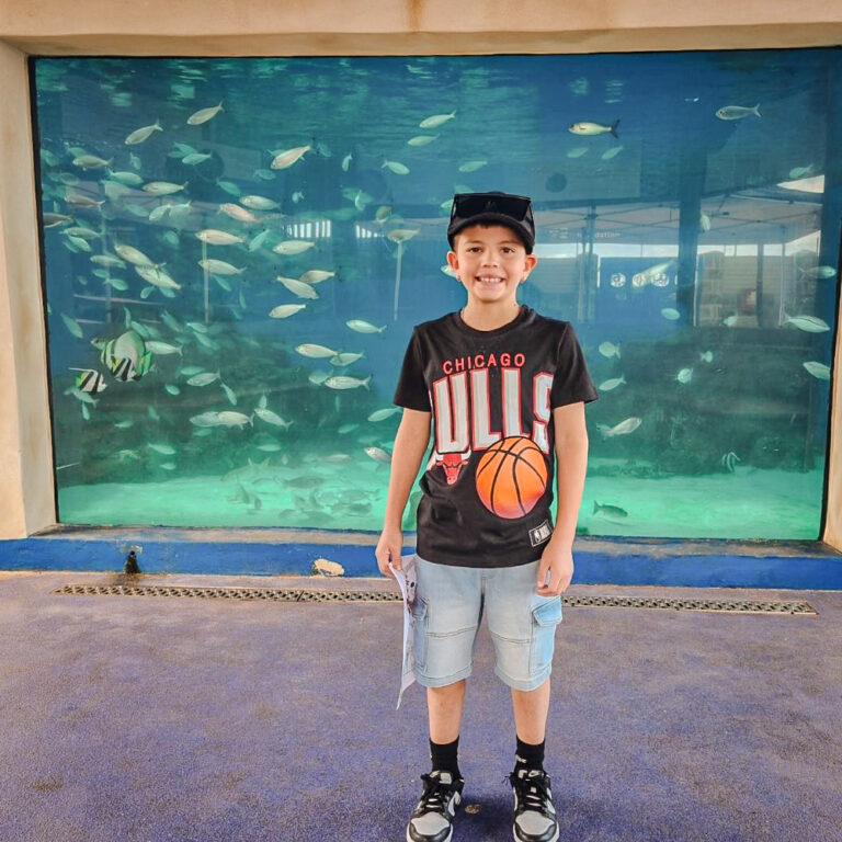 A young boy smiles while standing in front of a large aquarium display filled with fish. He is wearing a black Chicago Bulls shirt, light blue shorts, and a cap.