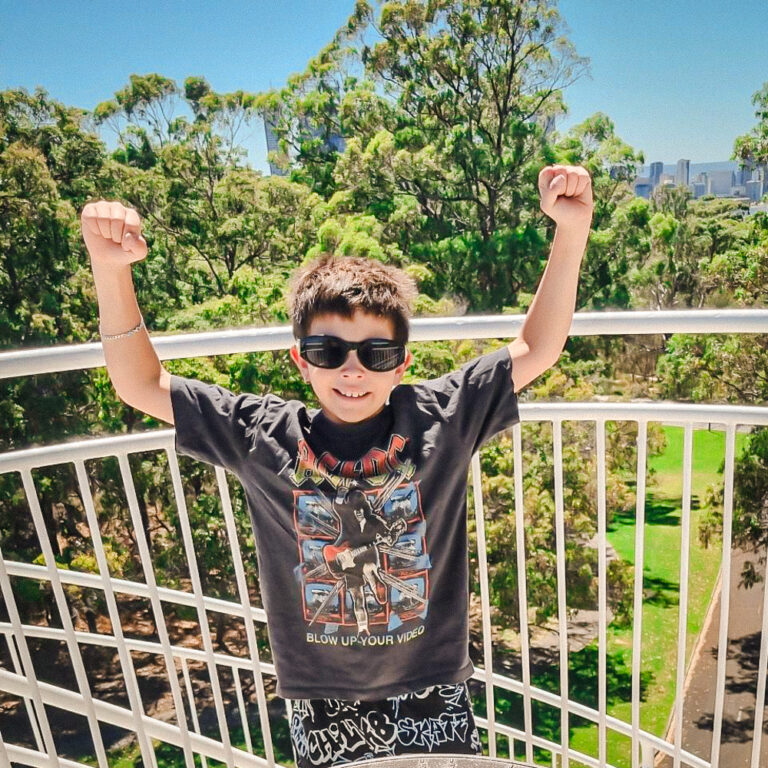 A young boy wearing sunglasses and a black AC/DC shirt stands on a balcony with both arms raised in a victory pose. He is smiling, with green trees and a city skyline in the background.