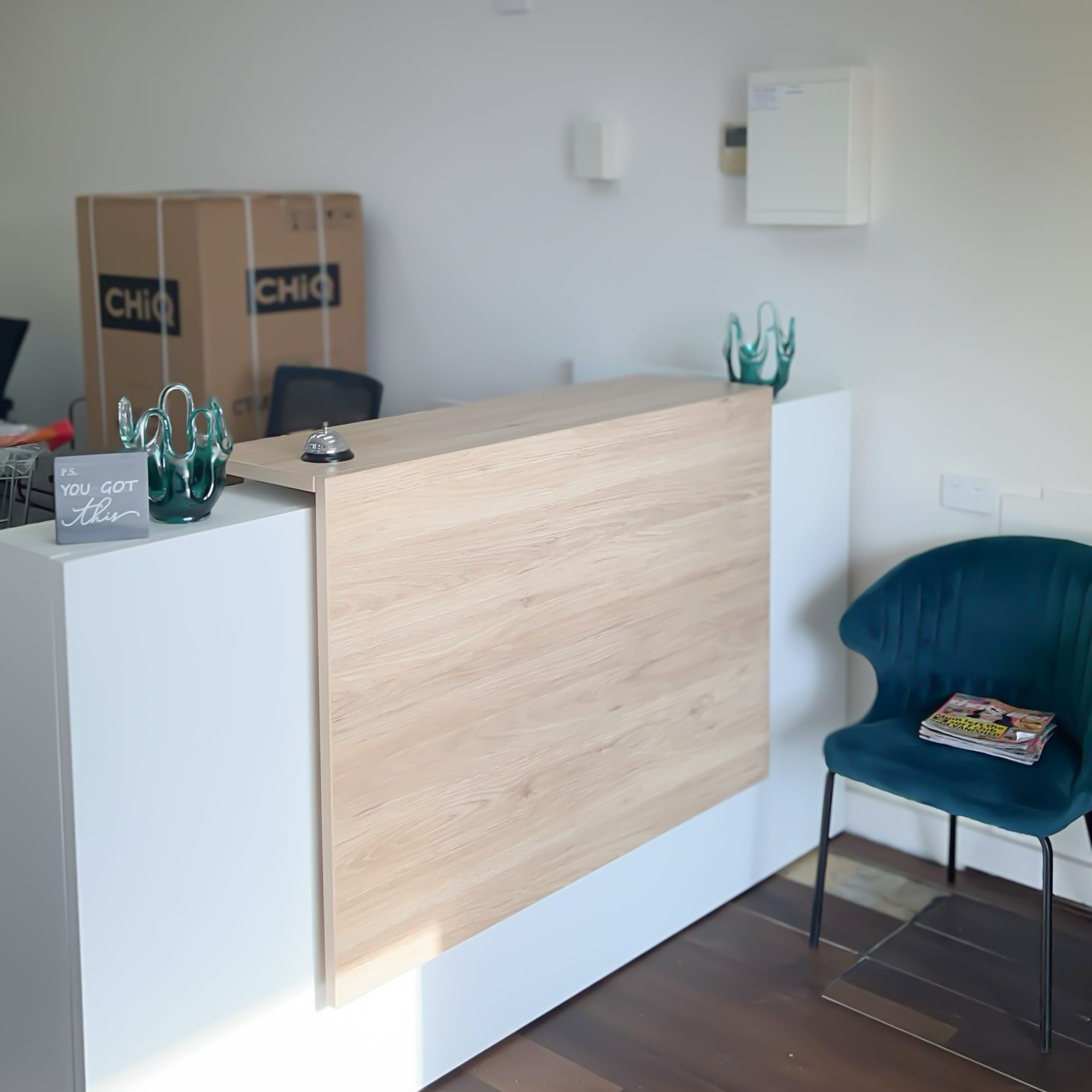 A close view of the reception desk at the Together Now WA office in Ballajura, with a light timber-fronted counter, a service bell, a small "P.S. you got this" sign and decorative teal glass sculptures on the side counter, a Deep Teal velvet armchair with a few magazines on the seat to the right of the desk, and the rest of the office space visible behind the counter.