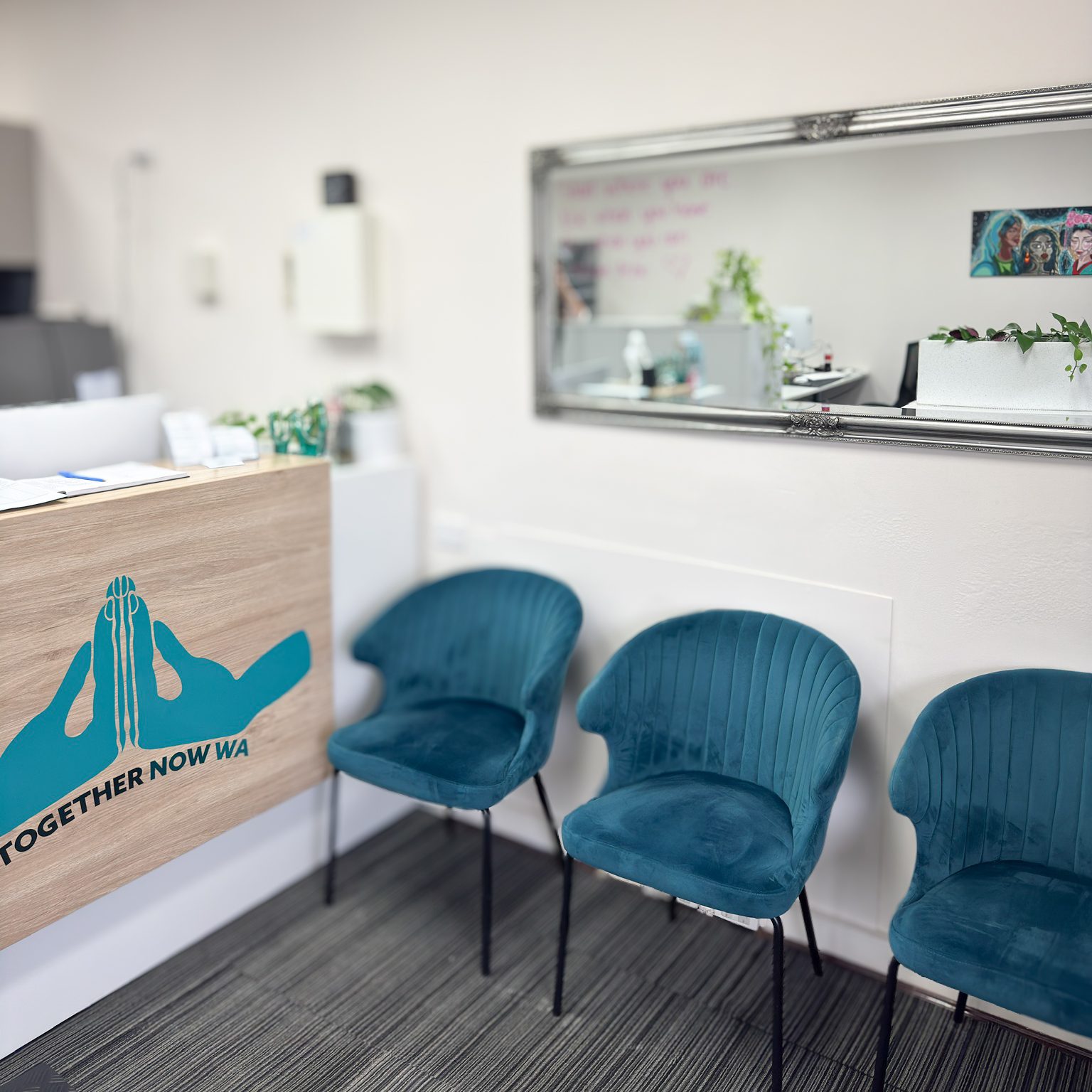 The reception waiting area inside the Together Now WA office in Ballajura, with a wooden-fronted reception desk on the left carrying a large Together Now WA hands logo and wordmark, three Deep Teal velvet upholstered armchairs lined up along the white wall on the right, a wide gilt-framed mirror above the chairs reflecting the office behind, indoor plants on the reception bench, and a small framed artwork on the wall.