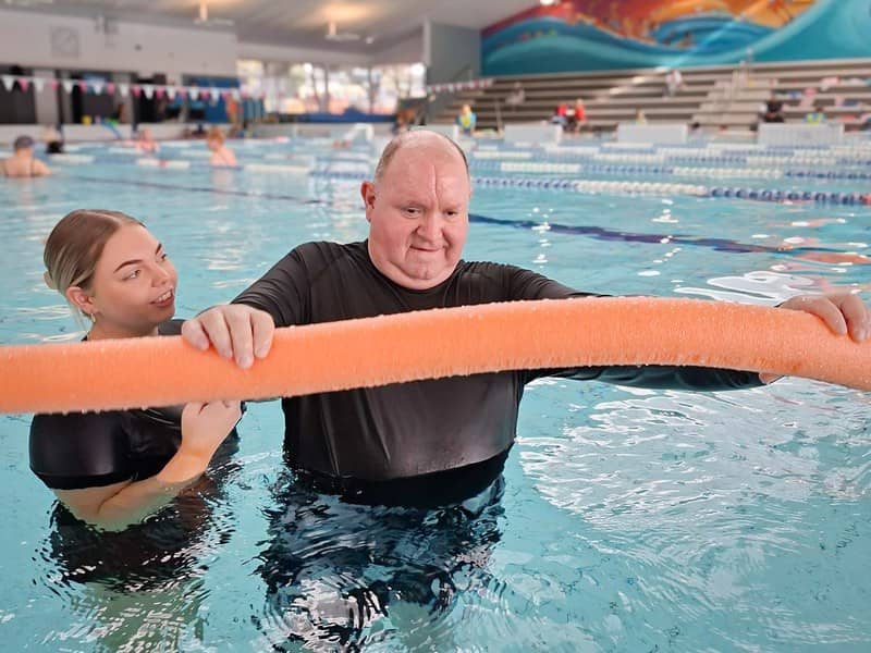 A physiotherapist stands in a public swimming pool beside a participant, both holding an orange pool noodle during a supported aquatic session.