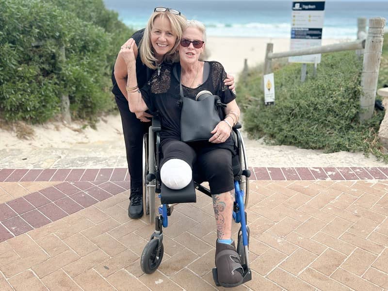 A TNWA support worker leans down beside a participant in a wheelchair at a Perth beach, both smiling, with the ocean in the background.