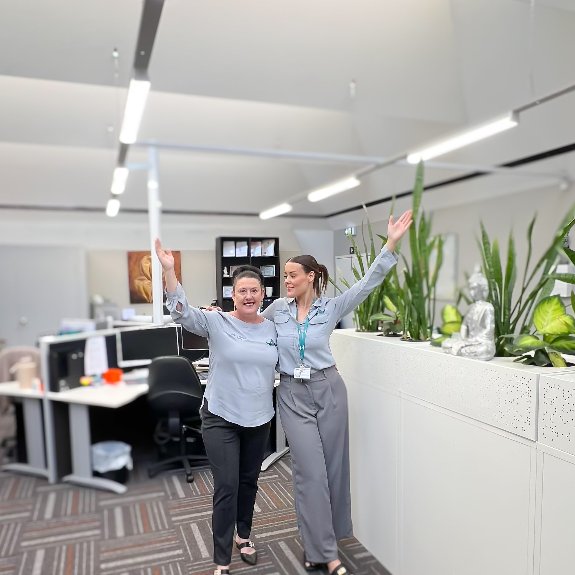 Two TNWA office staff in matching light blue Together Now WA polos standing arm in arm with both arms raised overhead, smiling toward the camera in the Ballajura open-plan office, with desks, indoor plants, and a small Buddha statue on a benchtop visible in the background.