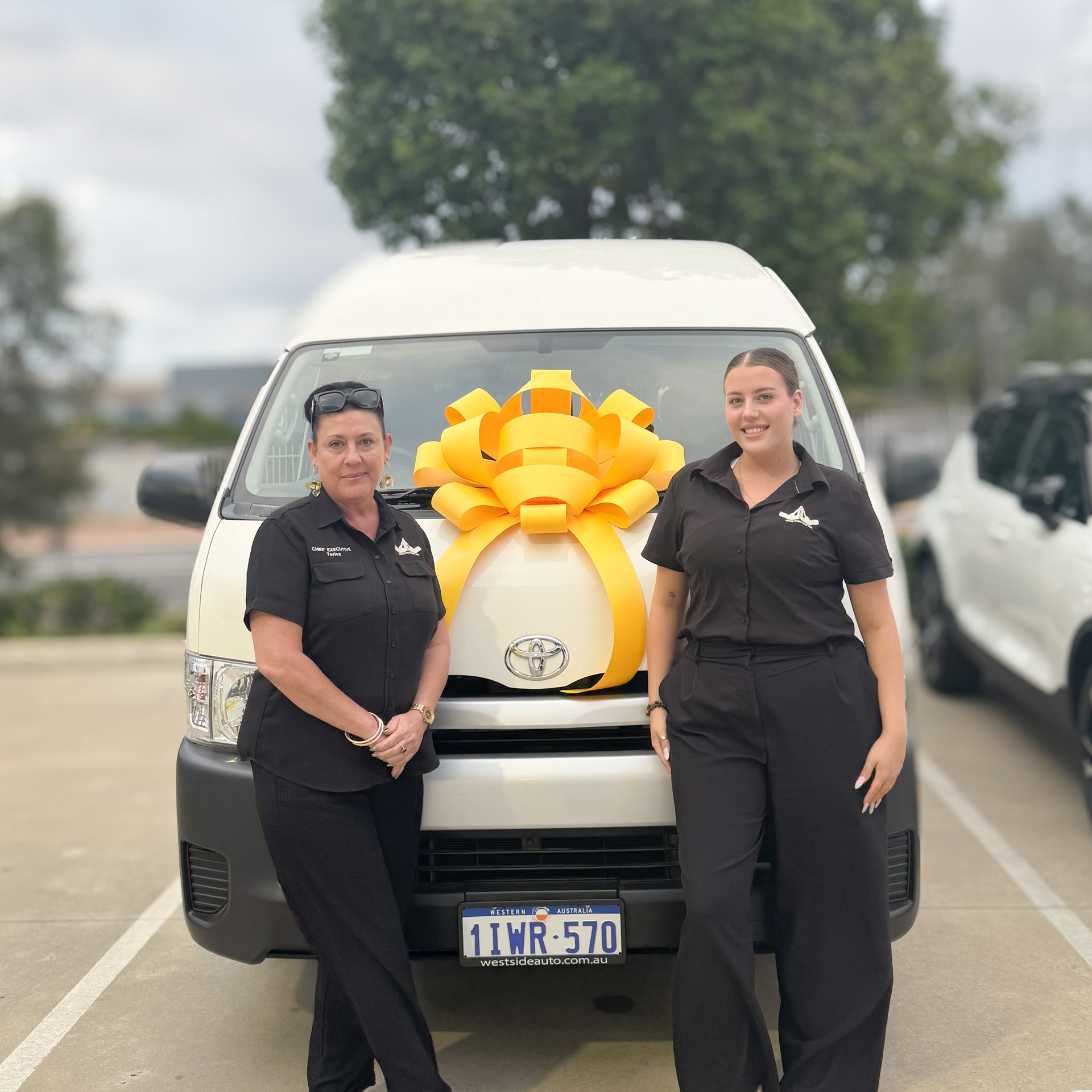 TNWA founder and Chief Executive Terica Isgar in a black Chief Executive polo standing on the left of a new white Toyota Hiace community transport van, alongside a TNWA staff member in a black Together Now WA polo on the right, both smiling toward the camera, with a large yellow gift bow on the van's bonnet and a Western Australian number plate visible.
